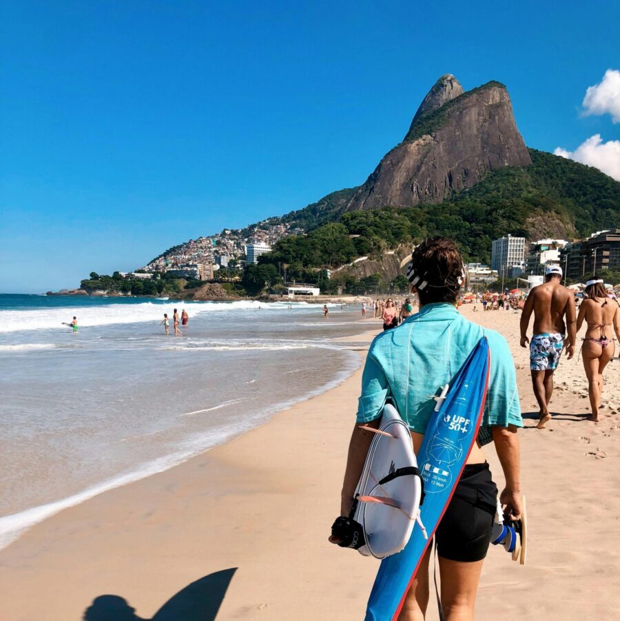 A surfer carrying a board strolls down Ipanema Beach, Rio de Janeiro, under a sunny sky, with a scenic mountain backdrop.